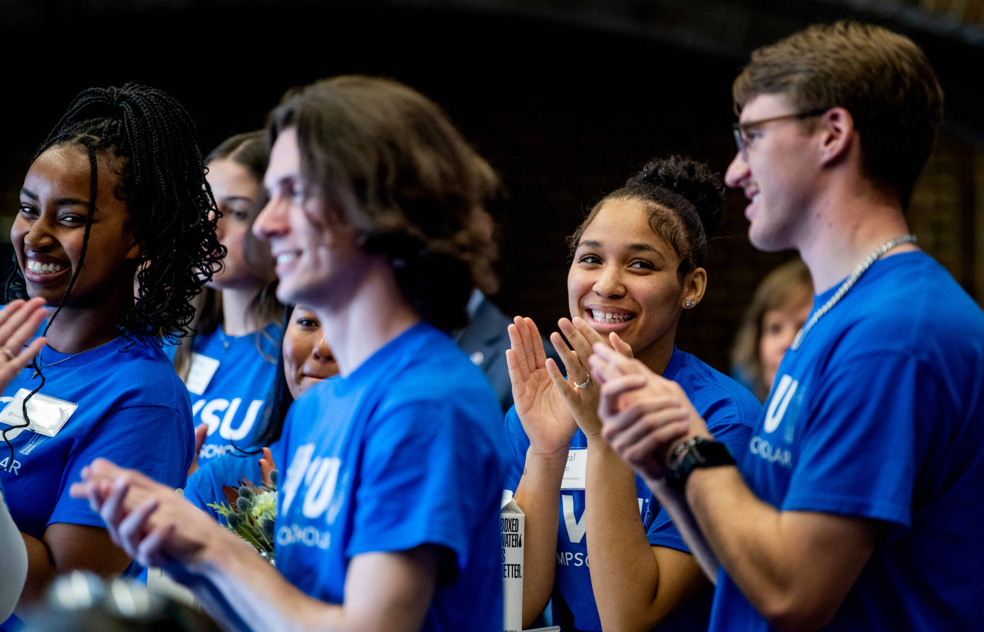 A group of students wearing blue Thompson Scholar t-shirts are seen clapping and smiling.